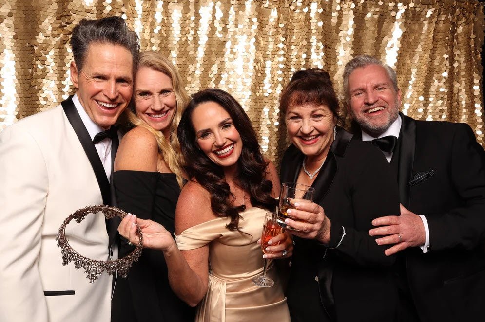 Group of five adults dressed in formal attire at a celebration, standing in front of a gold sequin backdrop, smiling, and holding drinks.