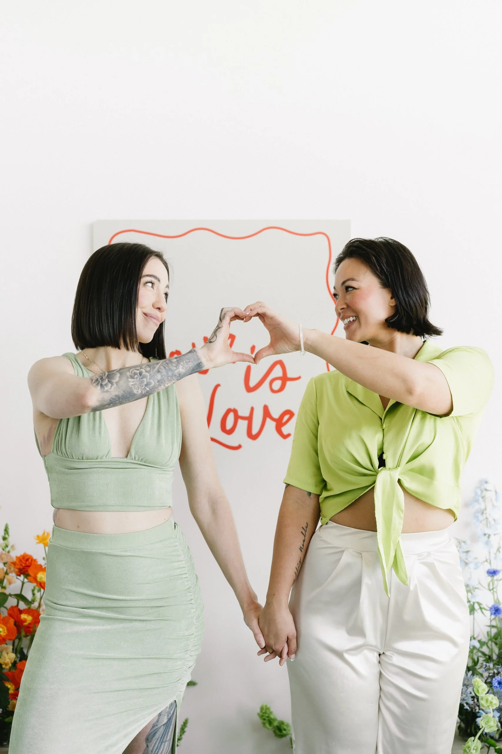 Two women are holding hands and forming a heart shape with their arms. They are standing in front of a white wall with a red and white sign that says 'love'. The woman on the left has dark hair, tattoos on her arm, and is wearing a light green dress. The woman on the right has short dark hair, and is wearing a lime green top and white pants. There are colorful flowers at the bottom of the image.
