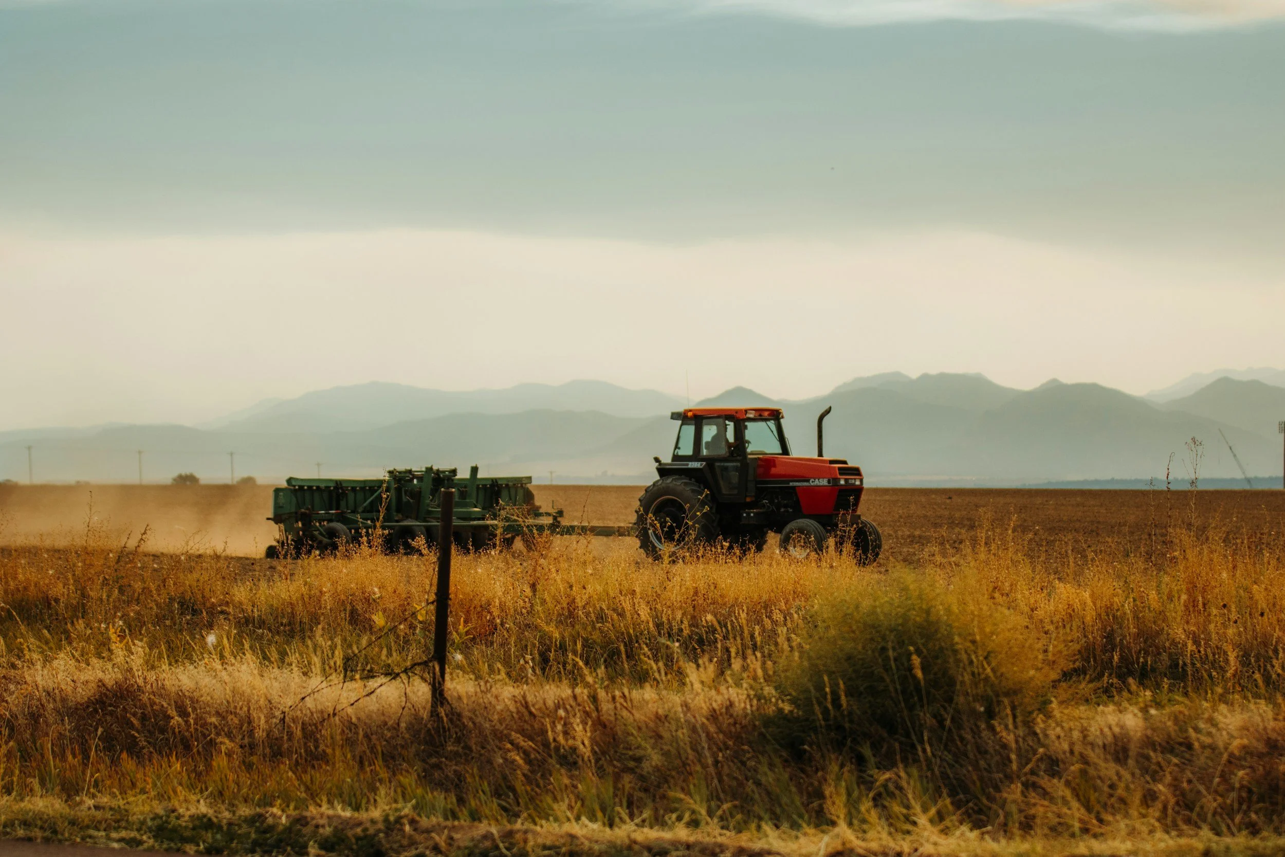 Red tractor working on a dry, yellow field with mountains in the background under a cloudy sky.