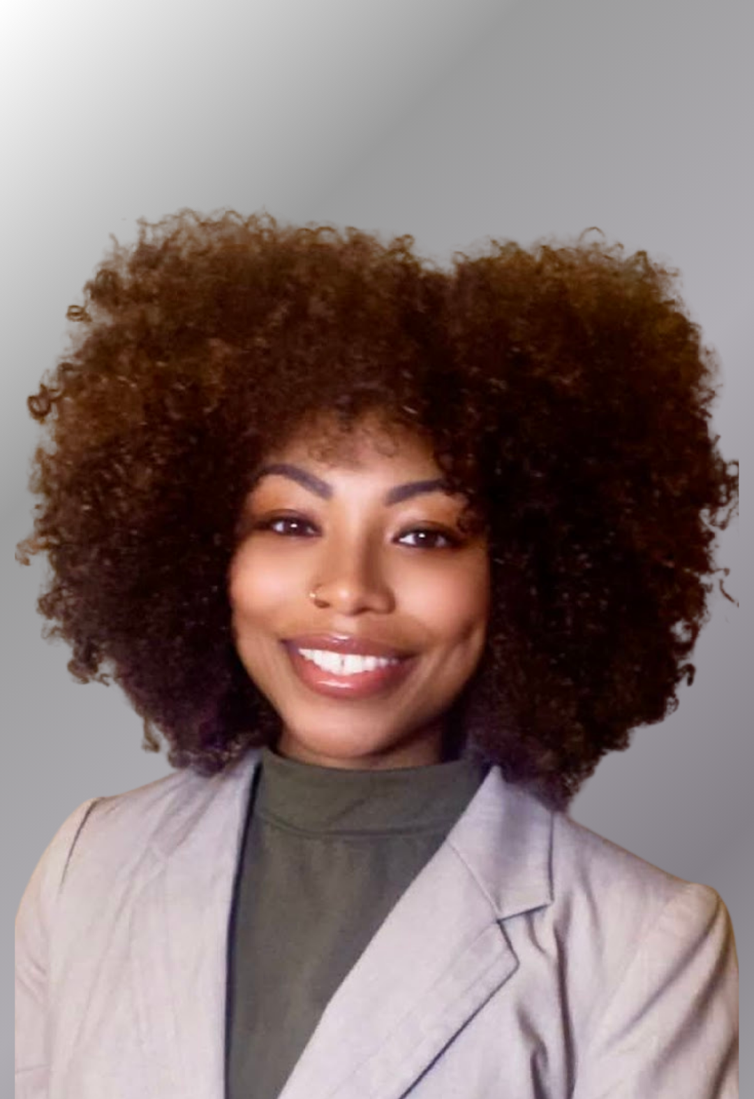 A woman with curly, voluminous hair smiling at the camera. She has a nose piercing and is wearing a light blazer over a dark top, with a neutral gray background.
