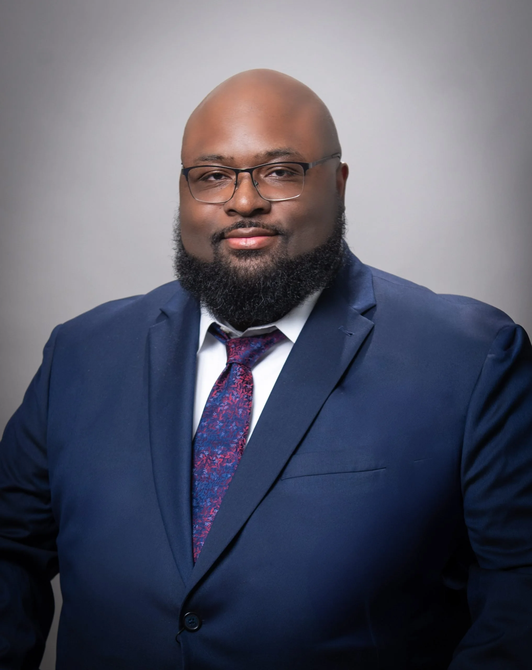 A man with a beard wearing glasses, a navy suit, white shirt, and patterned tie posing against a plain gray background.