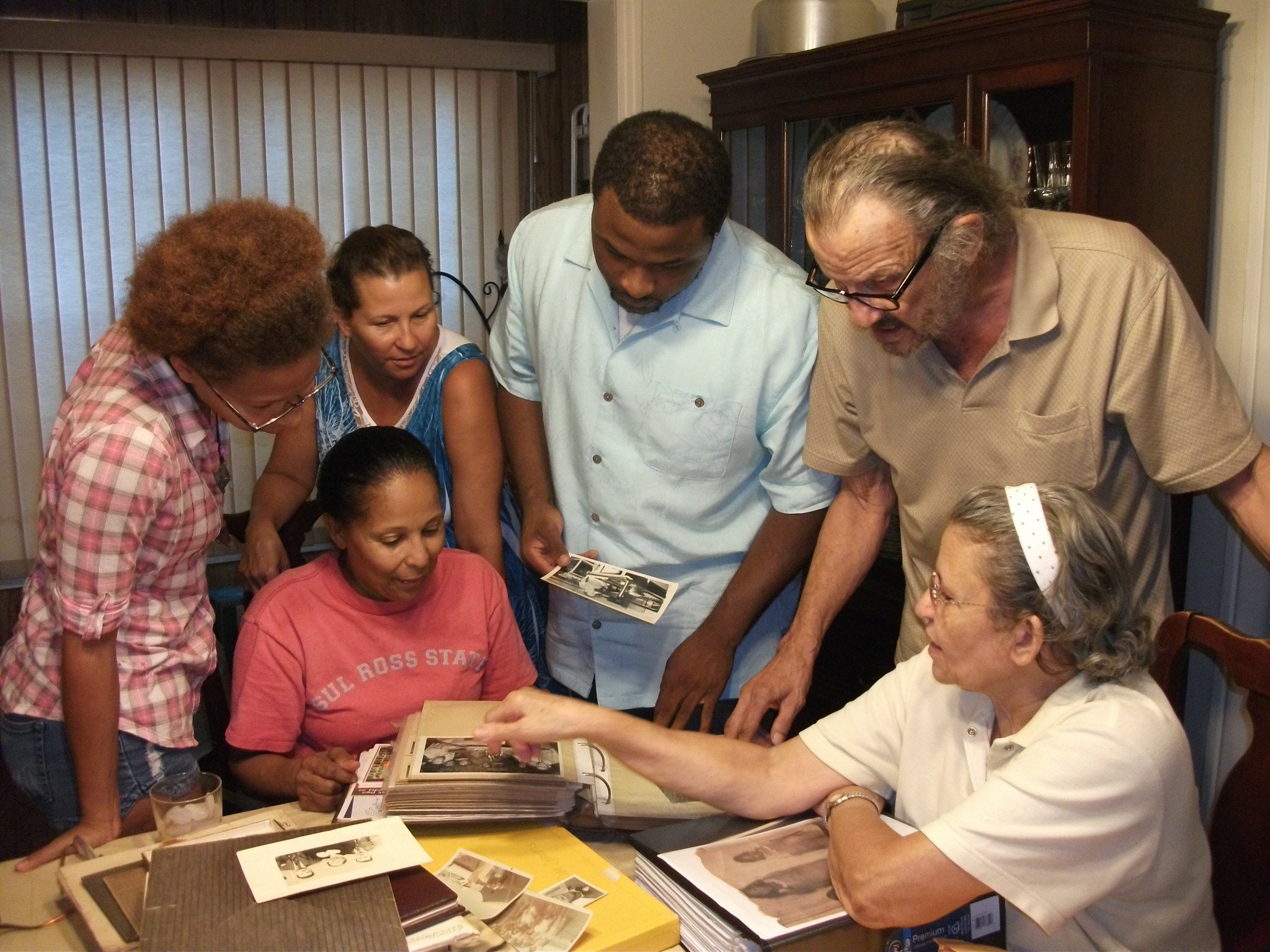 A group of seven people gathered around a table looking at photographs in photo albums, engaging in a discussion.