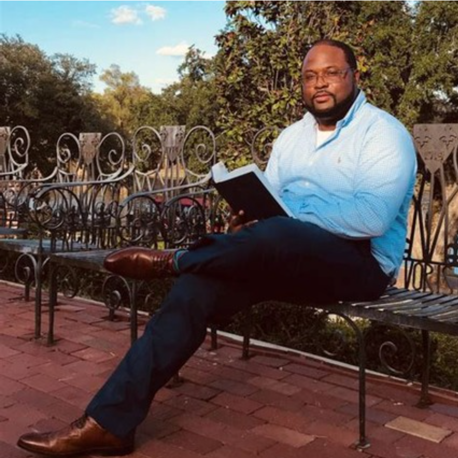 A man with glasses and a beard sitting on a park bench, holding an open book, outdoors during daytime with trees in the background.