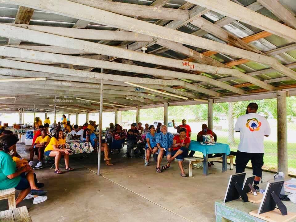 A group of people sitting under a wooden pavilion, listening to a person standing in front of them giving a presentation, with some tables and computer screens nearby.