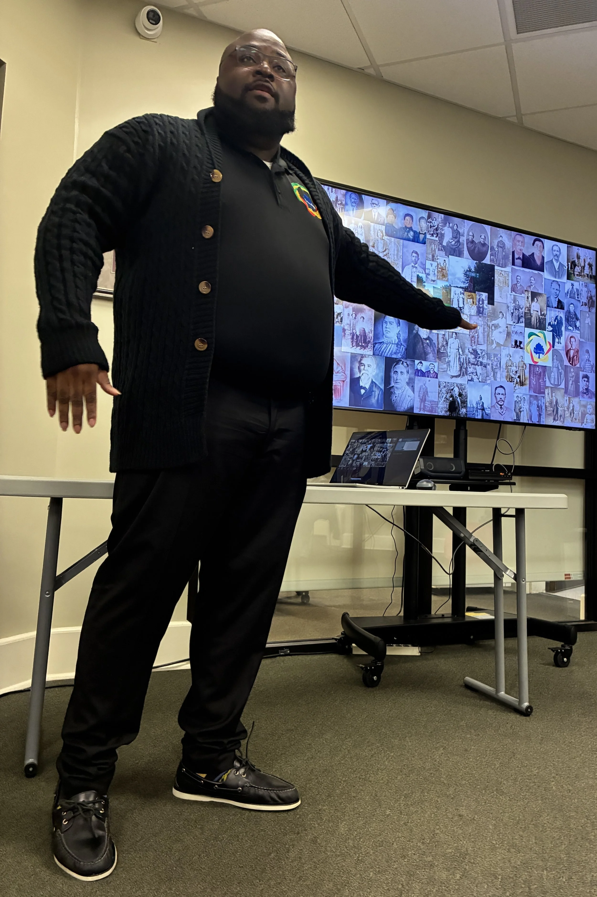 A man in black clothing pointing at a screen displaying a collage of black and white photos of various people.