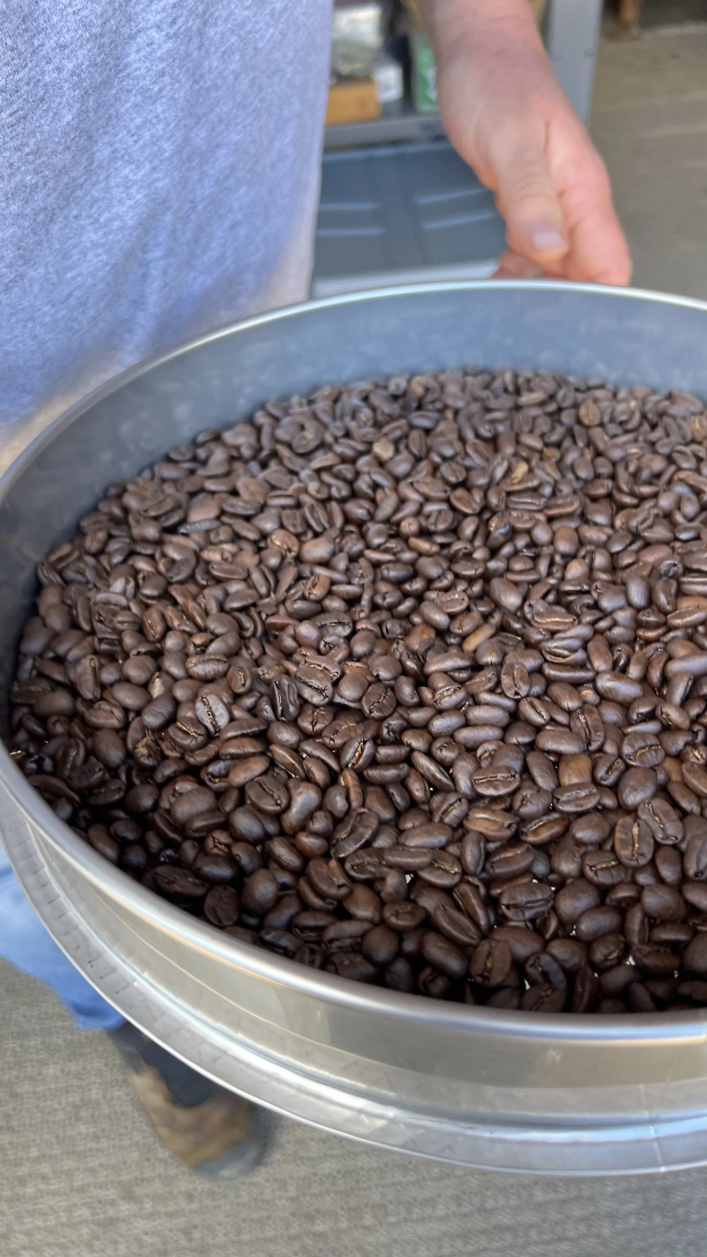 A person holding a large metal container filled with roasted coffee beans.