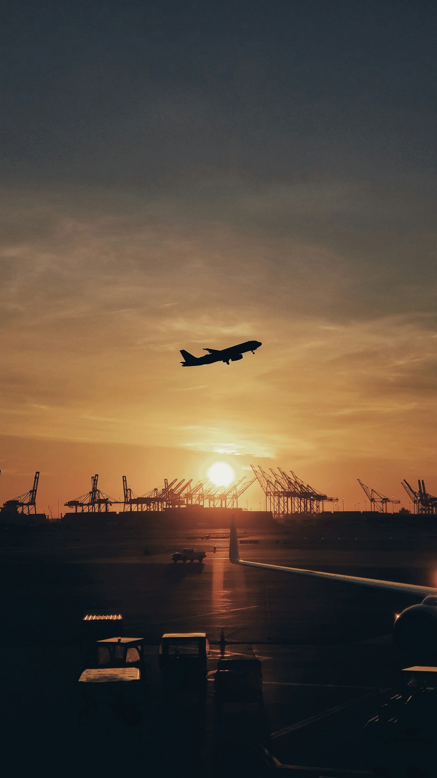 Silhouette of an airplane flying at sunset above an airport with cargo cranes in the background.