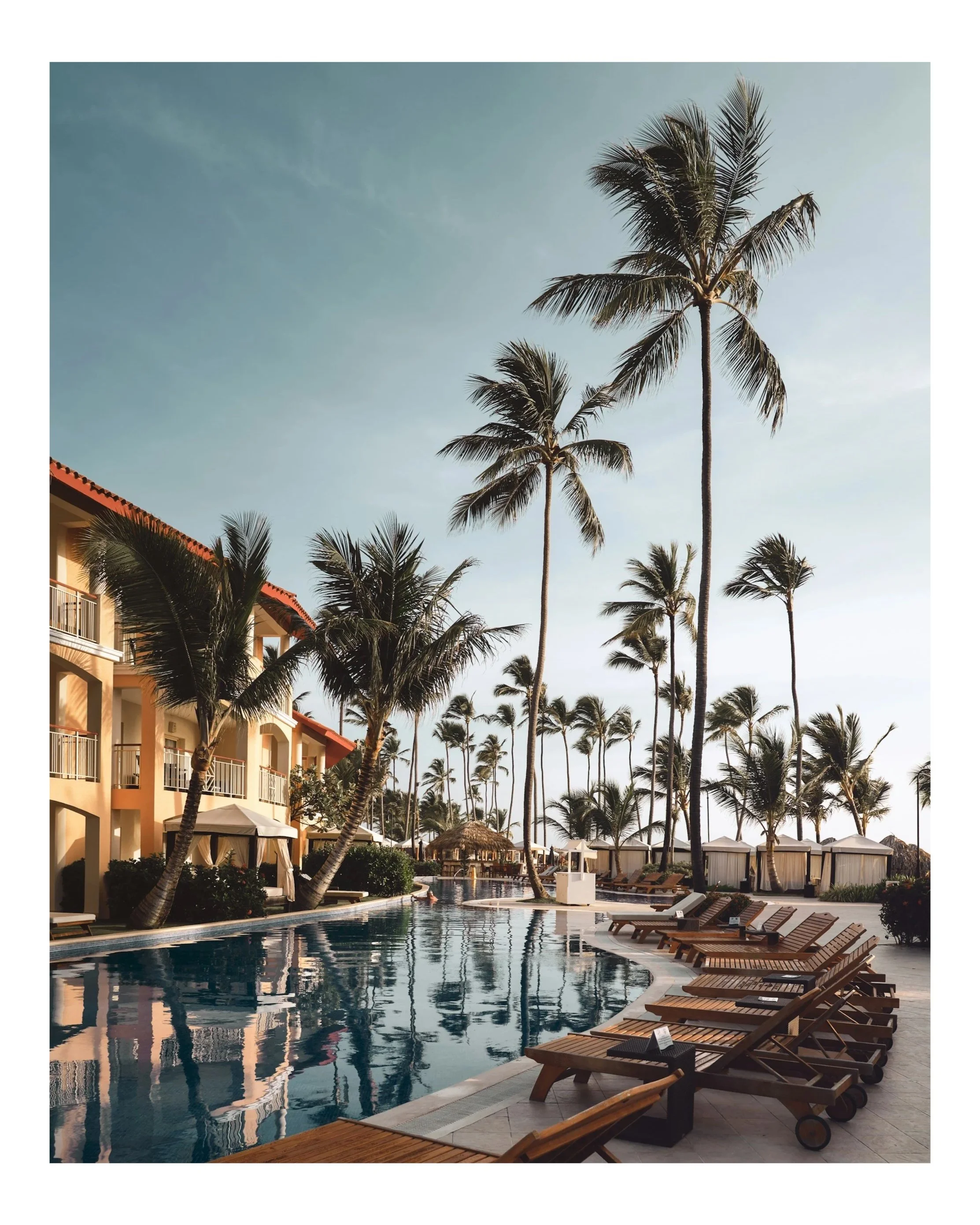 Poolside scene at a tropical resort with palm trees, lounge chairs, cabanas, and a building with balconies under a clear sky.
