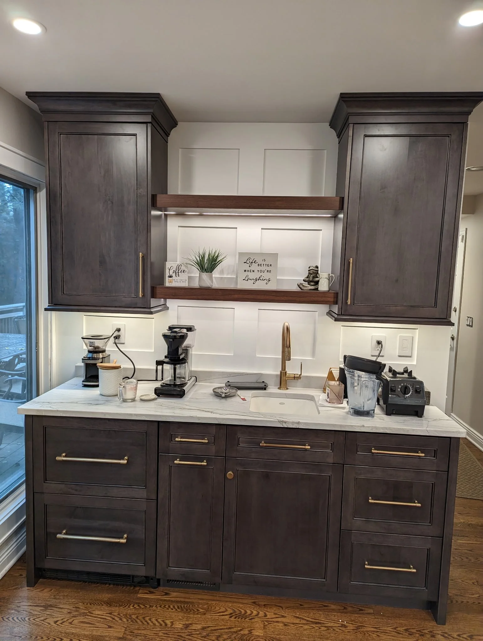 Kitchen with dark gray cabinets, marble countertop, and gold hardware, featuring coffee machines, a blender, and decorative items on open shelves.
