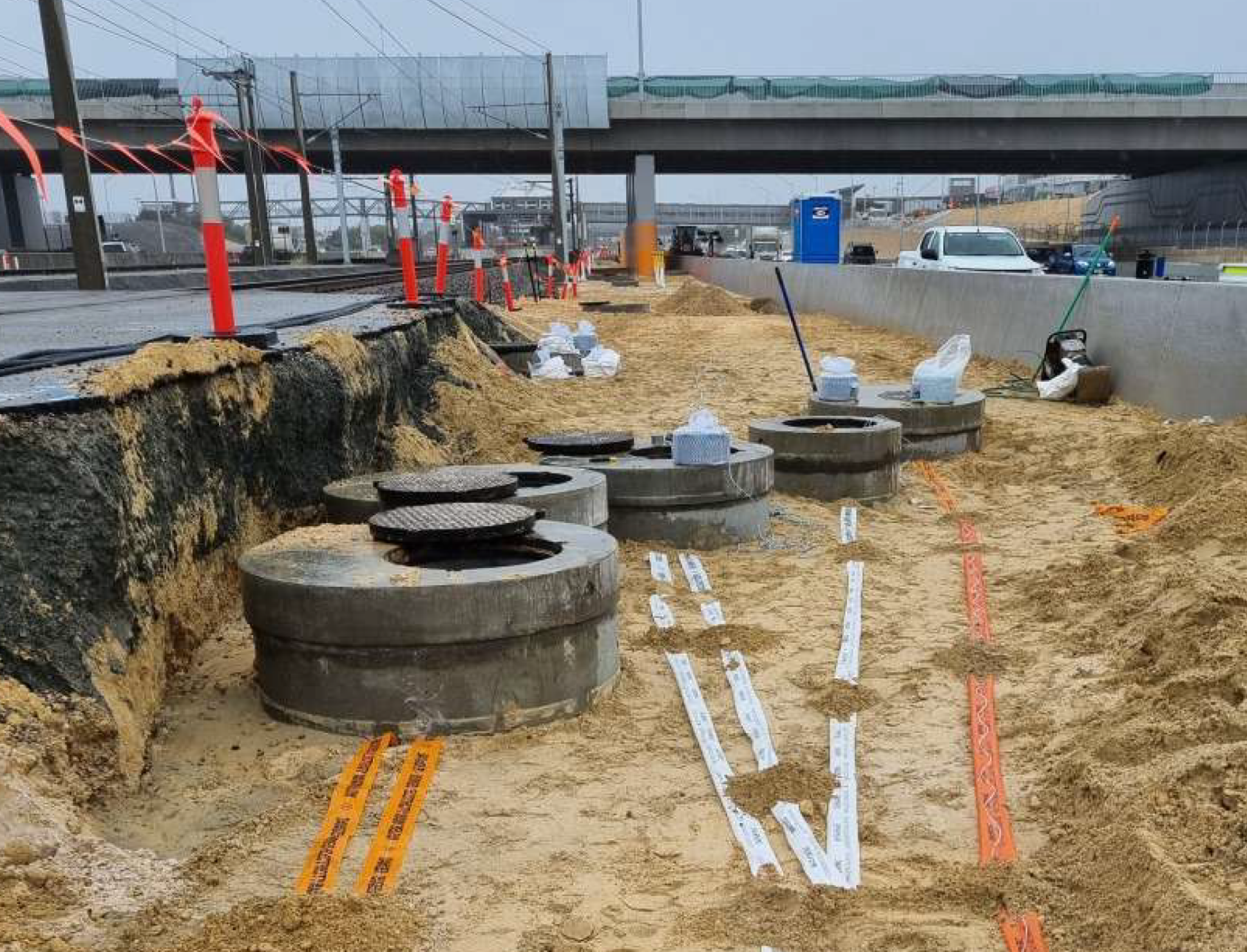Construction site showing large concrete pipes, orange safety barriers, and various equipment under a highway overpass.