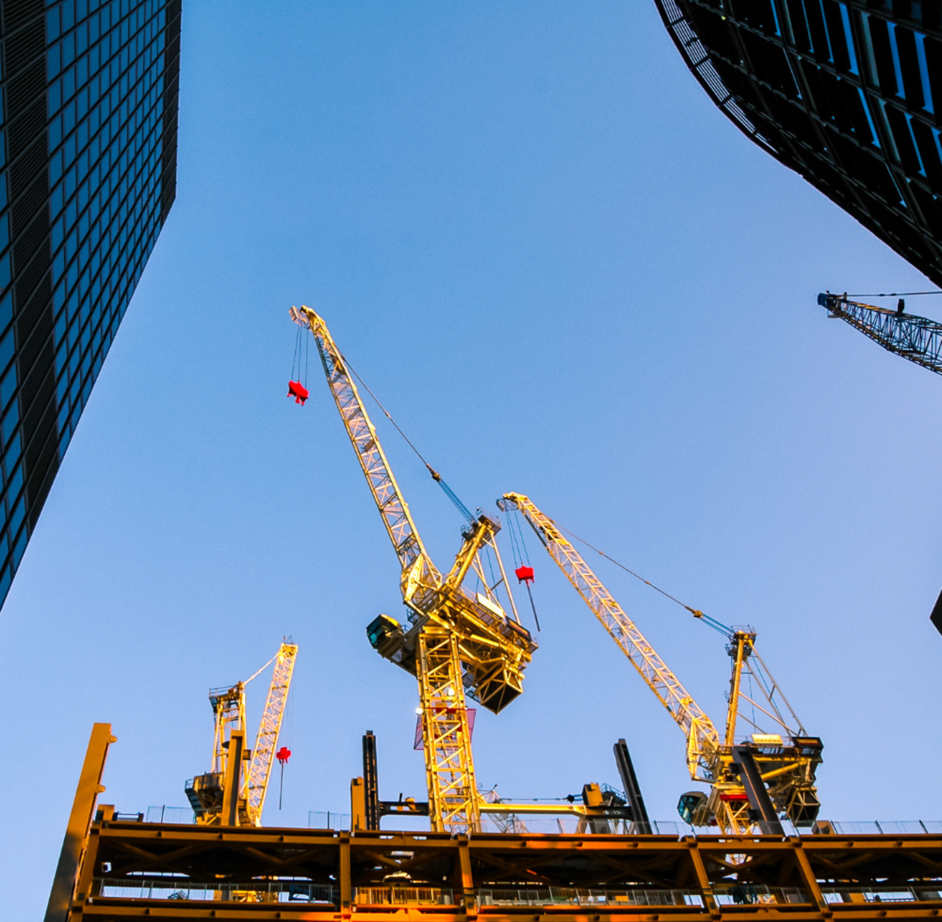 Construction site with multiple yellow cranes lifting materials against a clear blue sky, surrounded by tall buildings.