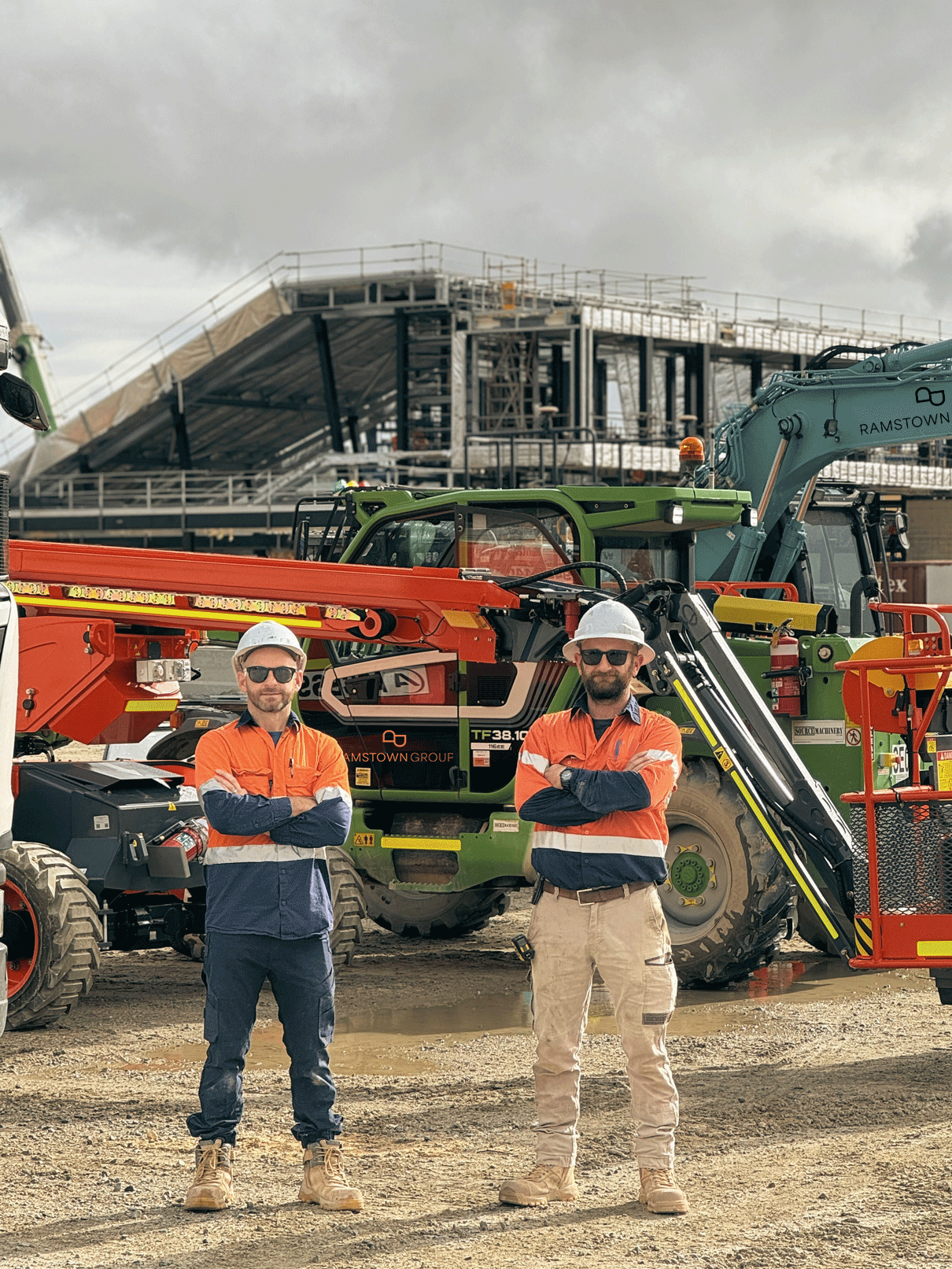 Two construction workers in safety gear, including helmets and sunglasses, standing with arms crossed in front of heavy construction machinery on a construction site with a partially built structure in the background under a cloudy sky.