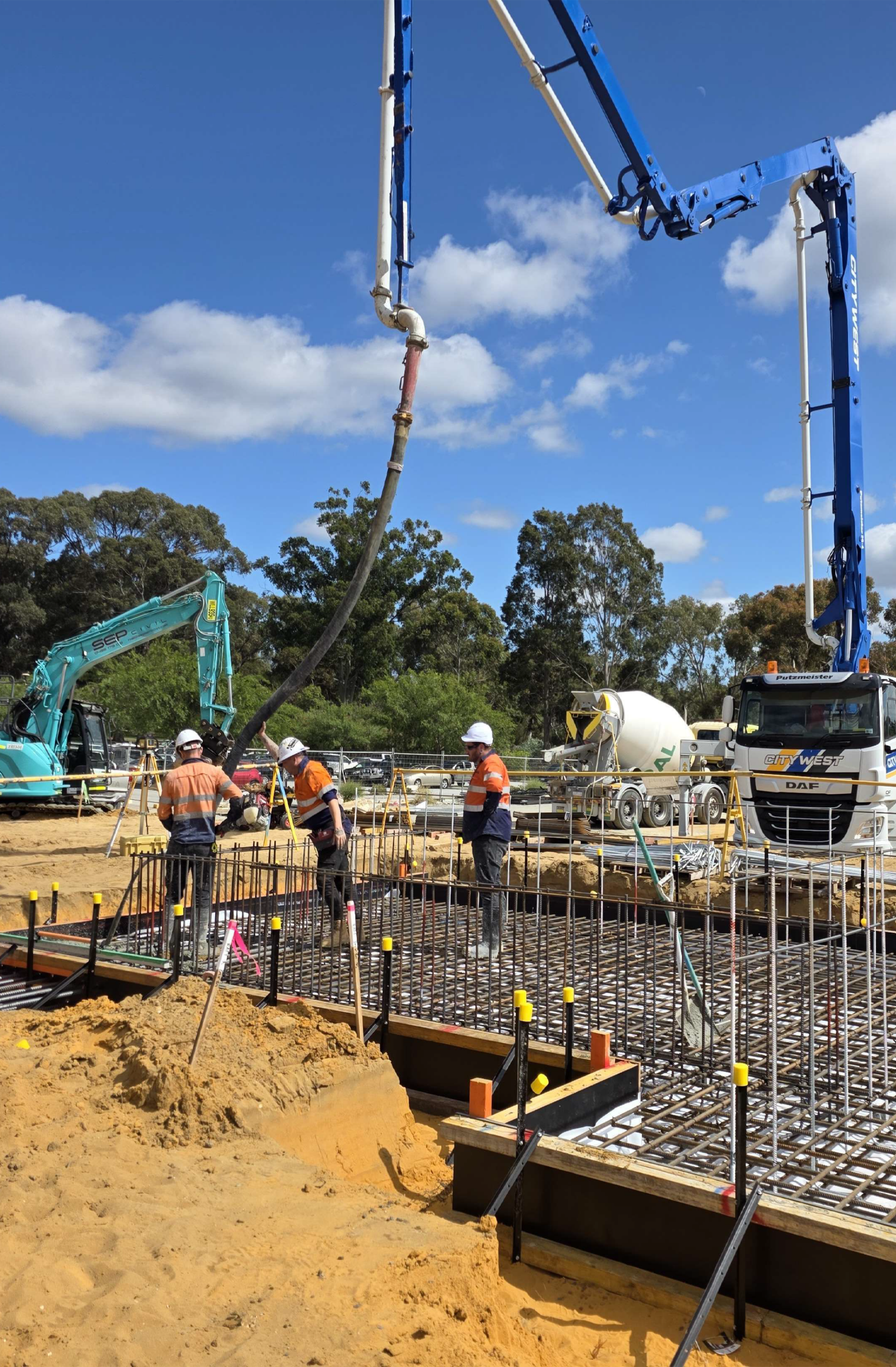Construction workers on a building site pouring concrete, with stabilizing rebar and construction machinery under a blue sky.