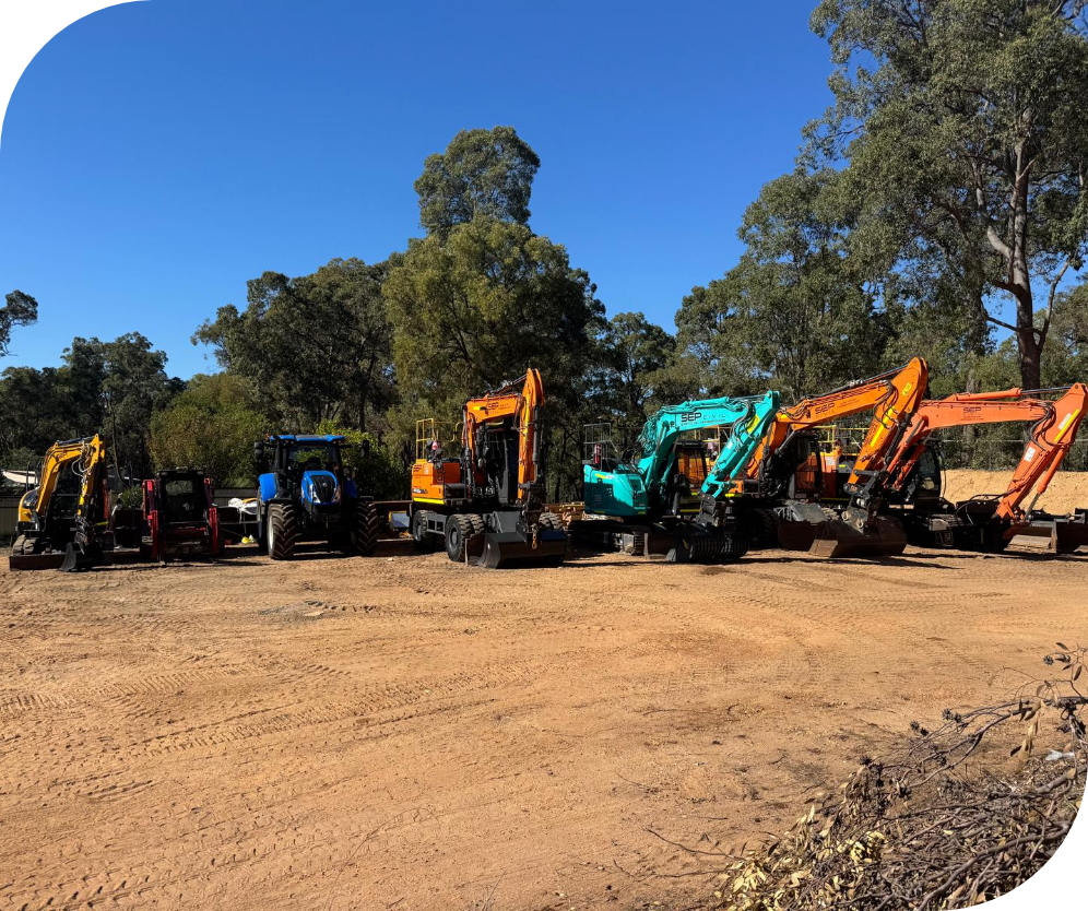 A row of construction excavators and tractors parked on a dirt lot with trees in the background and a clear blue sky.