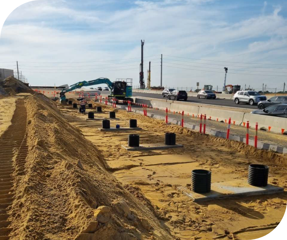 Construction site with asphalt pipes and equipment, cars driving by on an adjacent road under partly cloudy sky.