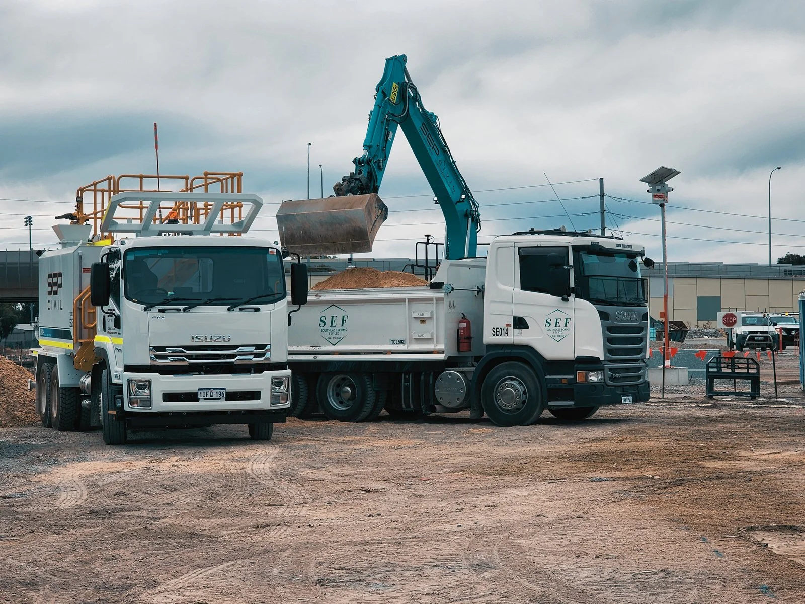 Construction site with two large trucks, one with a crane and excavator arm, working on dirt under cloudy sky, nearby street signs and fencing.