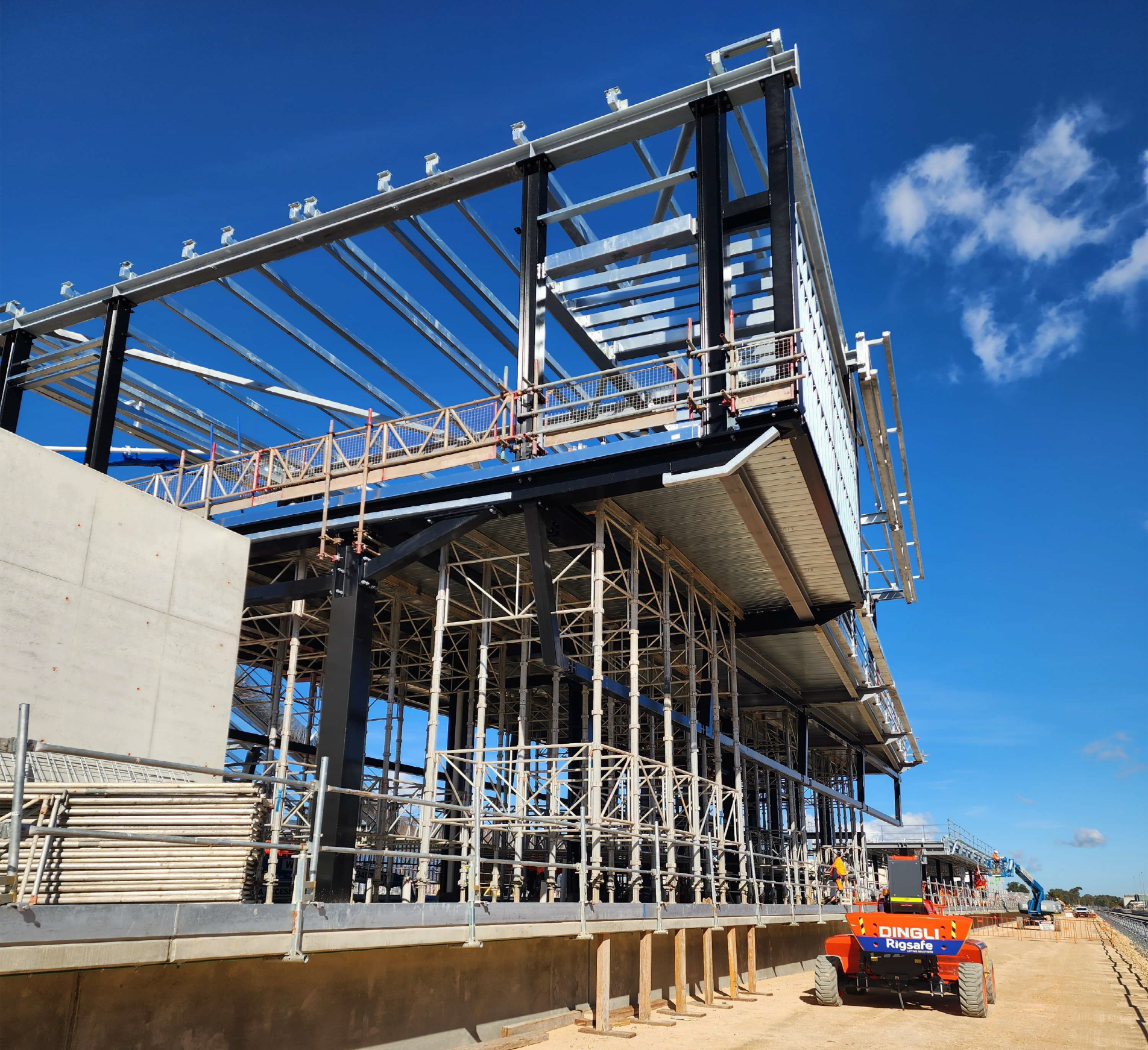 Construction site with scaffolding and building framework under a blue sky with few clouds, including a orange construction lift in the foreground.