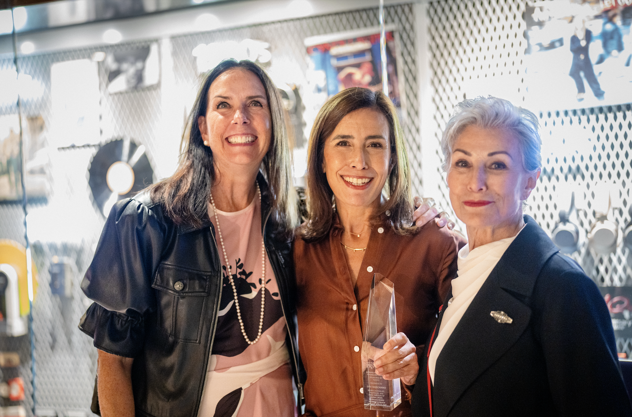Three women smiling, one holding a glass award, in an indoor setting with photos and artwork on the wall behind them.