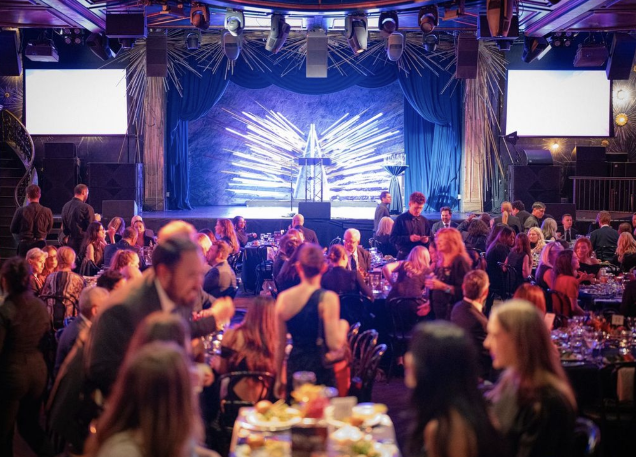 A large gathering of people in a dimly lit event hall with a decorated stage in the background, featuring blue curtains, lighting effects, and visual art installations, with tables set for dining and guests socializing.