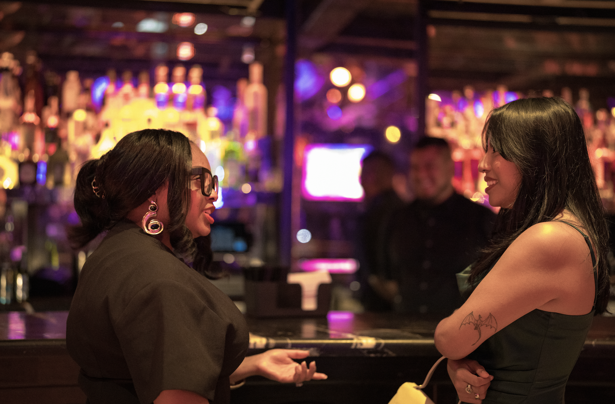 Two women having a conversation at a bar with colorful lights in the background, one with glasses and large earrings, the other with a tattoo on her arm and dark hair.