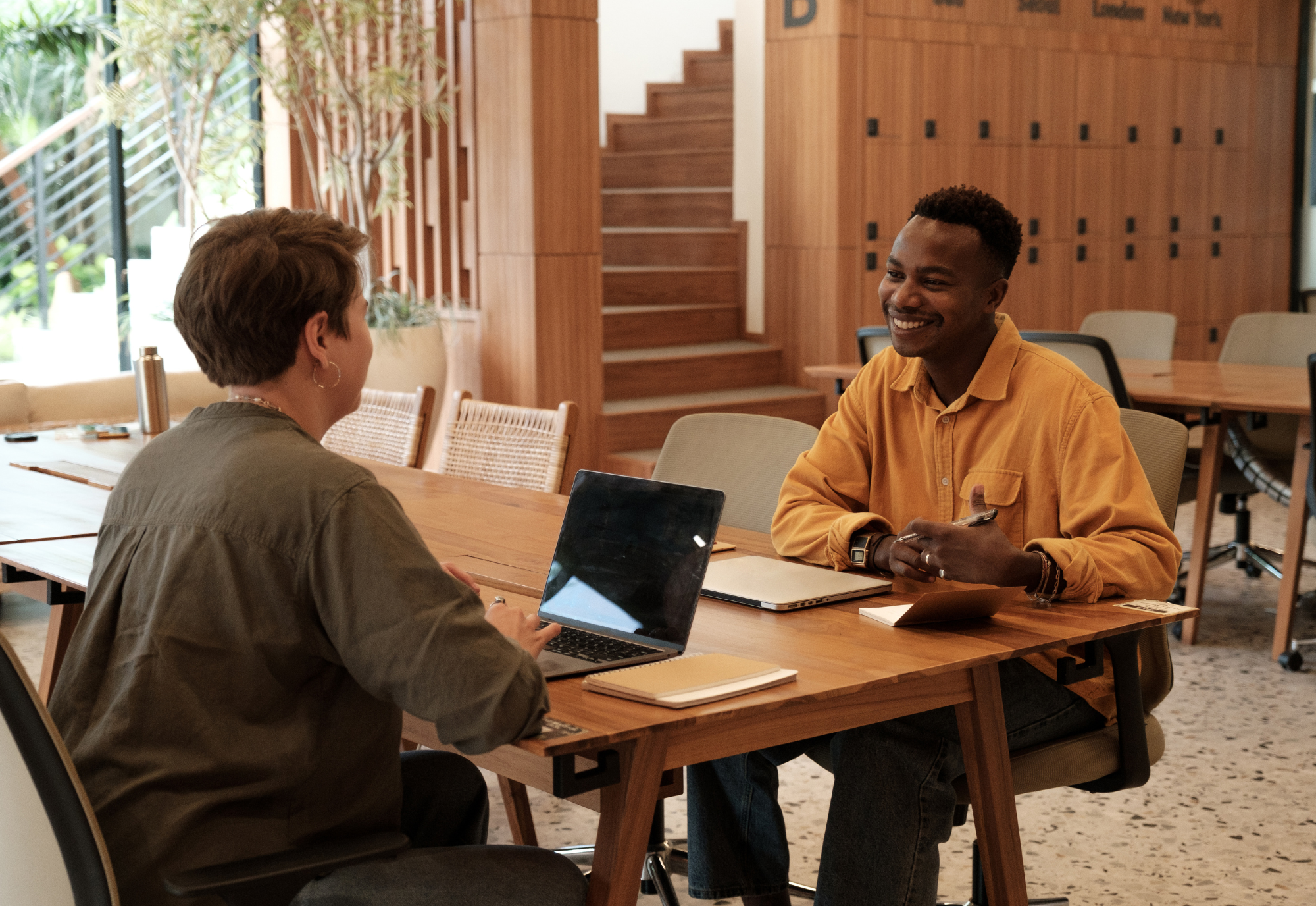 Two people sitting at a conference table having a conversation in a modern, wooden-furnished room. The person on the left has short brown hair, wearing a green shirt, and the person on the right has short black hair, wearing a yellow shirt, smiling, with a laptop, notebook, and tablet in front of them.