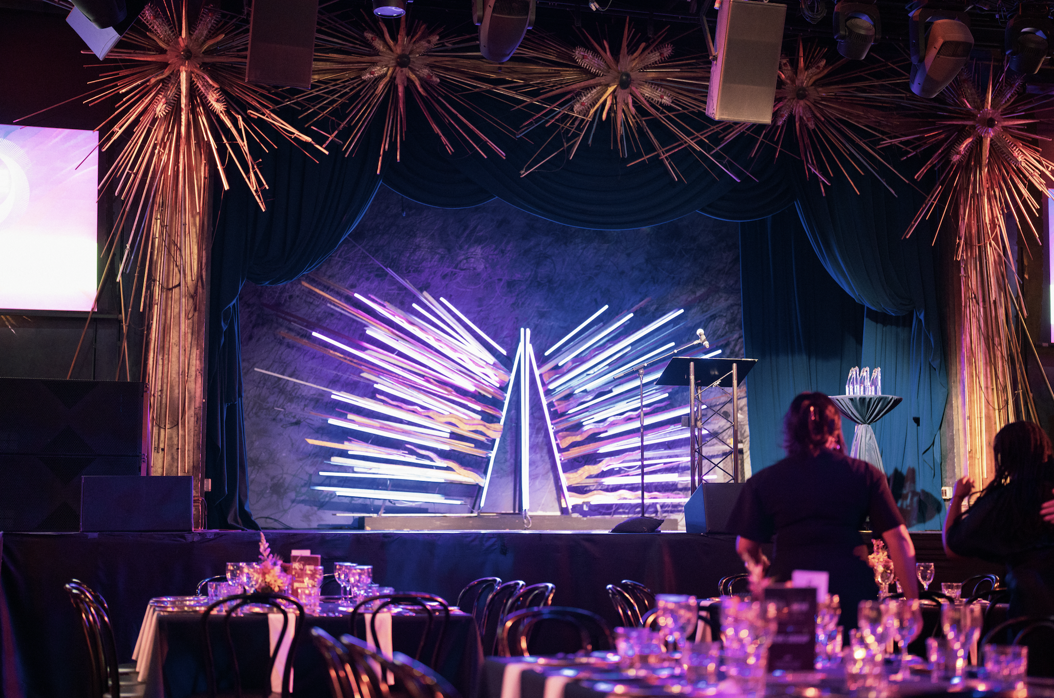 Indoor event space with a stage decorated with colorful lighting and neon lights, surrounded by blue curtains and gold starburst art. Tables with glassware and floral centerpieces are set up in the foreground, with some people standing near the stage.
