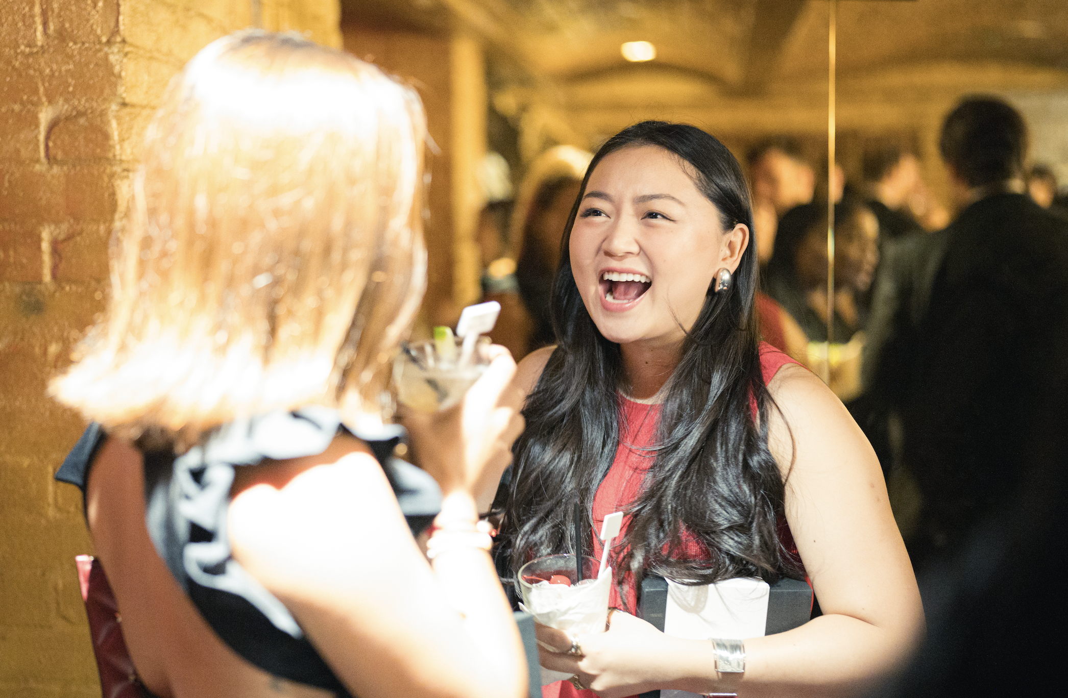 Two women having a lively conversation at a party, one is holding a drink and the other is smiling and laughing, with a crowded background.