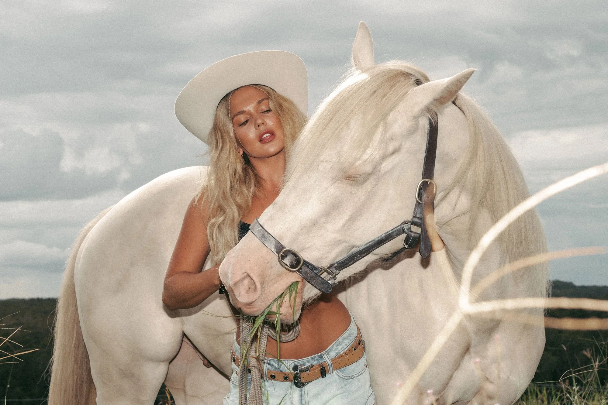 A woman with long blonde hair wearing a wide-brimmed hat and jeans stands beside a white horse in an open field with cloudy sky.