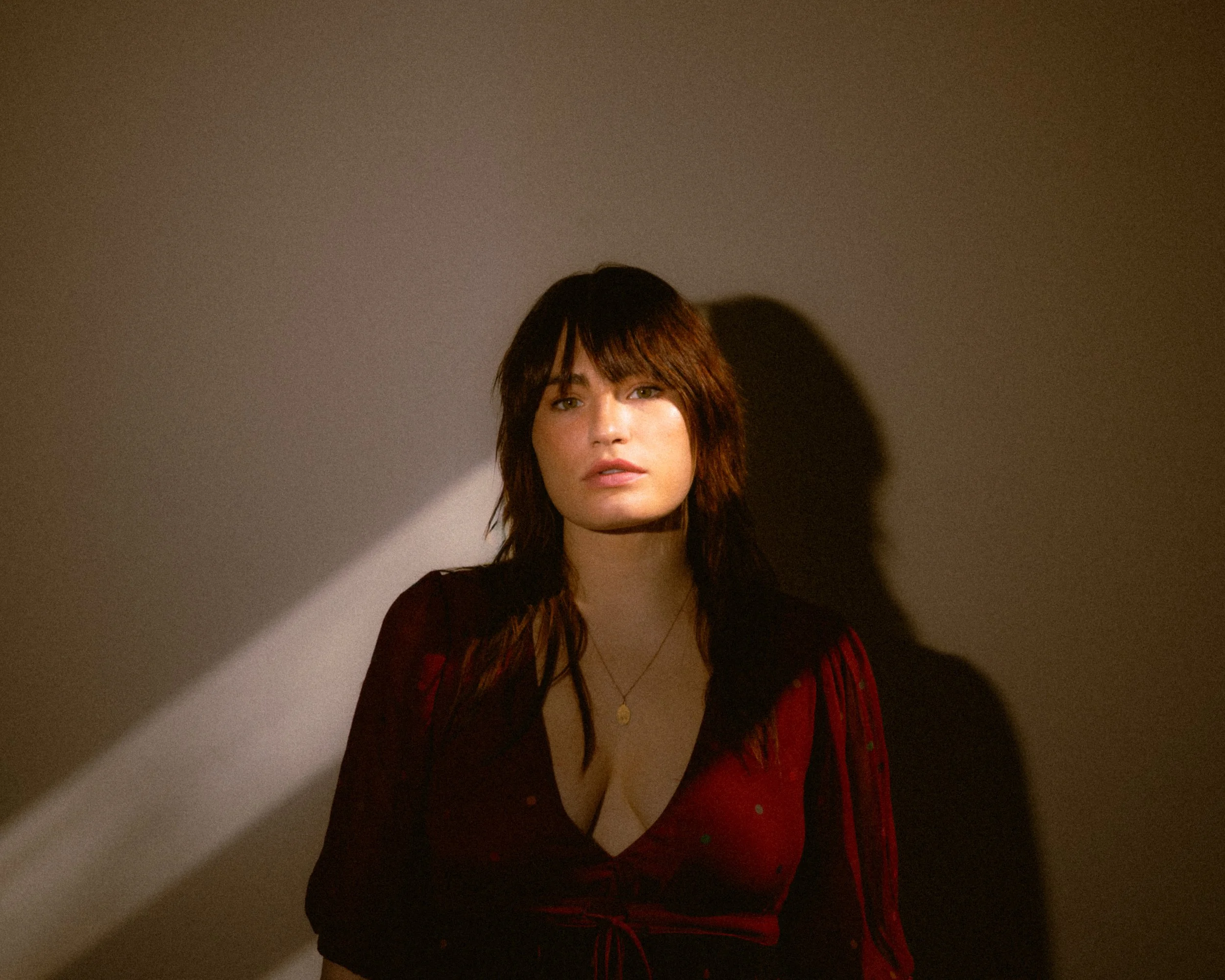 A woman with wavy brown hair and light skin sits against a plain wall, lit by a warm light creating a shadow behind her. She wears a deep red, low-cut top with small dots and a delicate gold necklace.