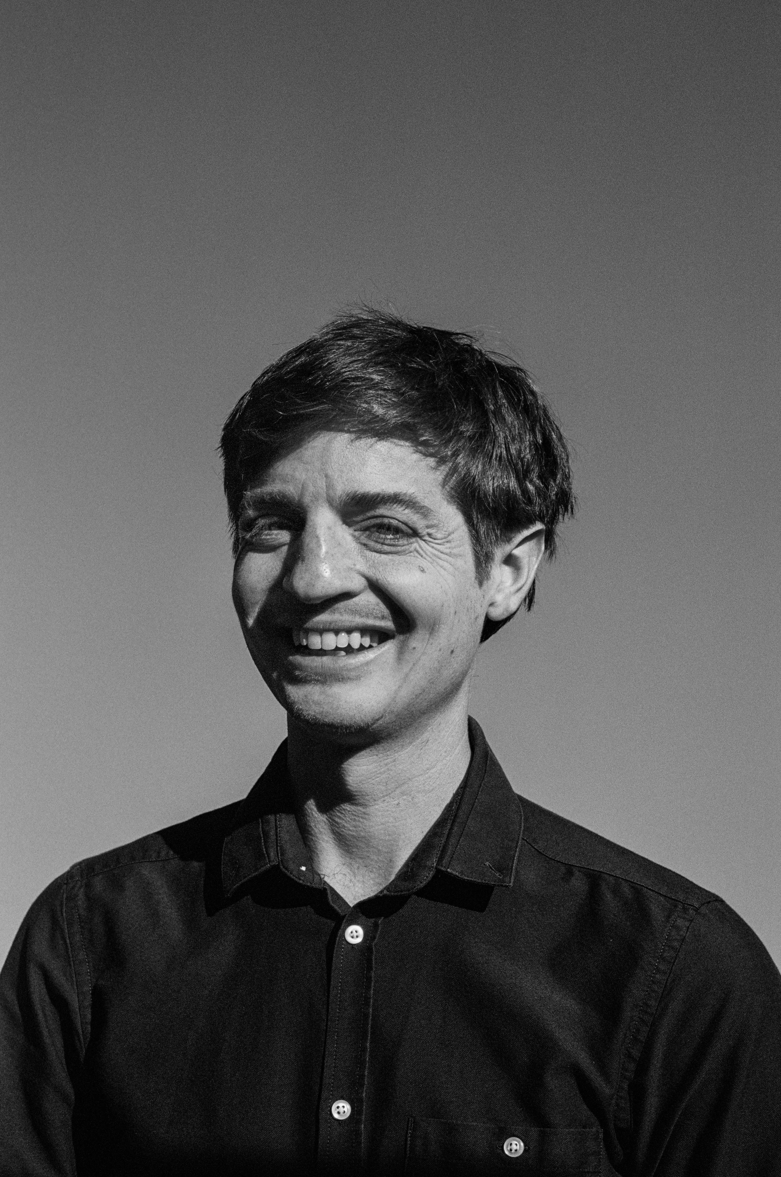 Black and white photo of a smiling man with short dark hair, wearing a button-up shirt, standing against a clear sky.