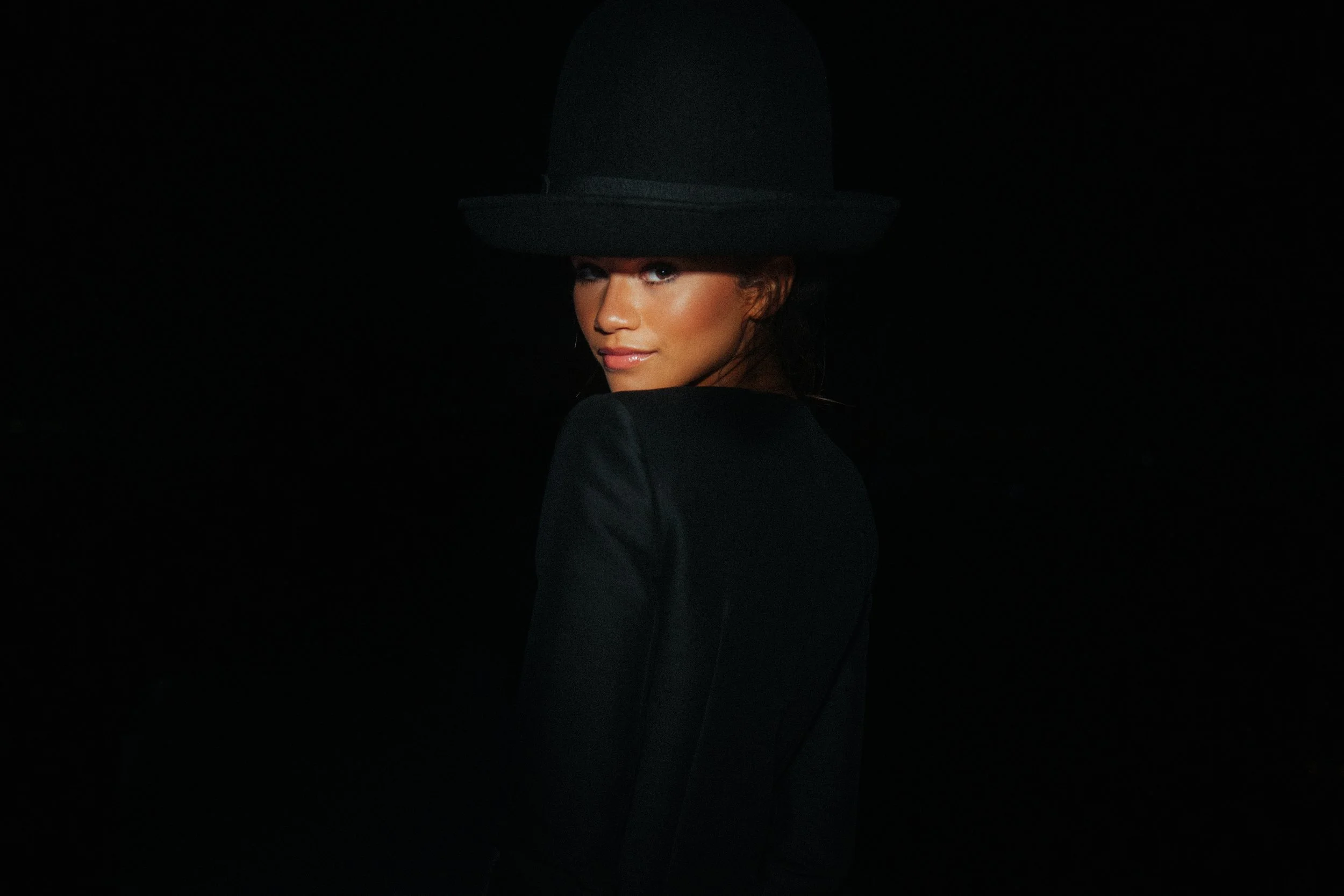 A woman in a black hat and black jacket looking over her shoulder against a dark background.