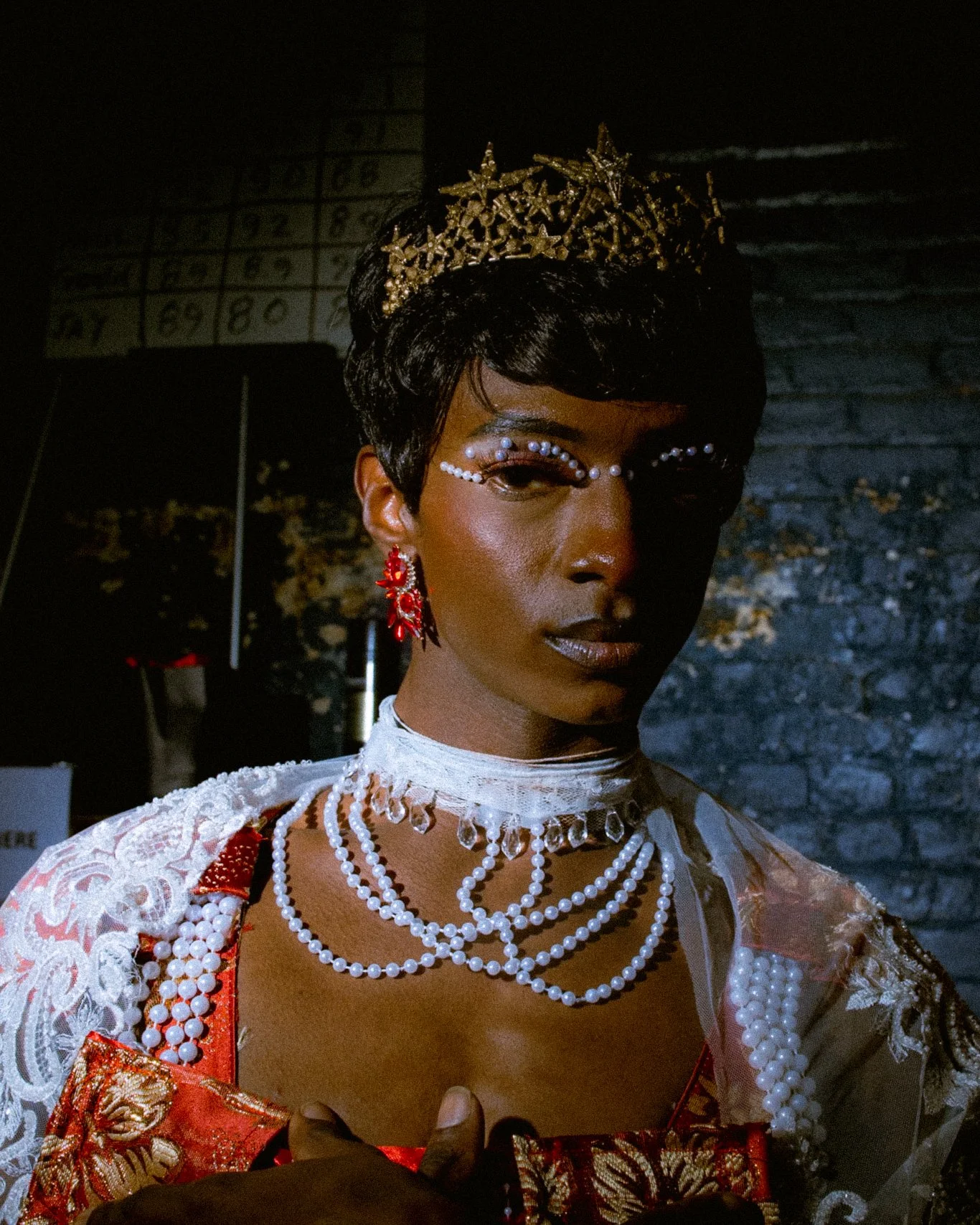 A person dressed in elaborate clothing and jewelry, wearing a crown, with makeup and accessories in a dark setting.