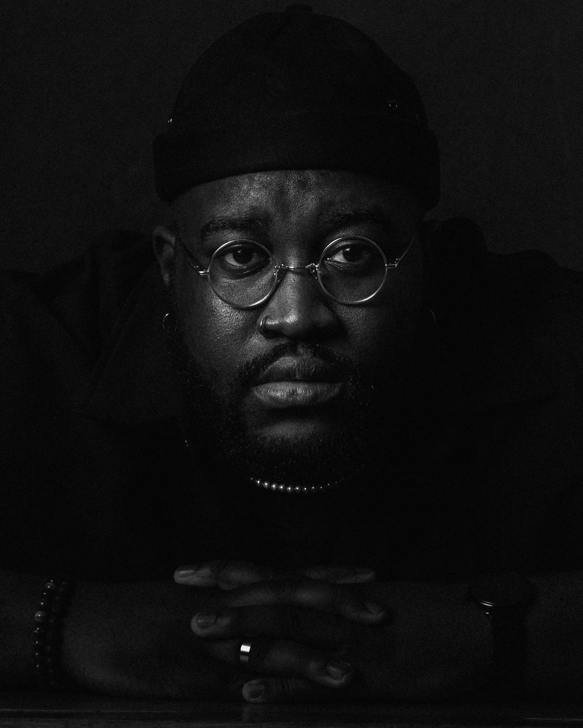 Black and white portrait of a man with glasses, a nose piercing, a beanie, and jewelry, looking directly at the camera with his hands clasped in front of him.