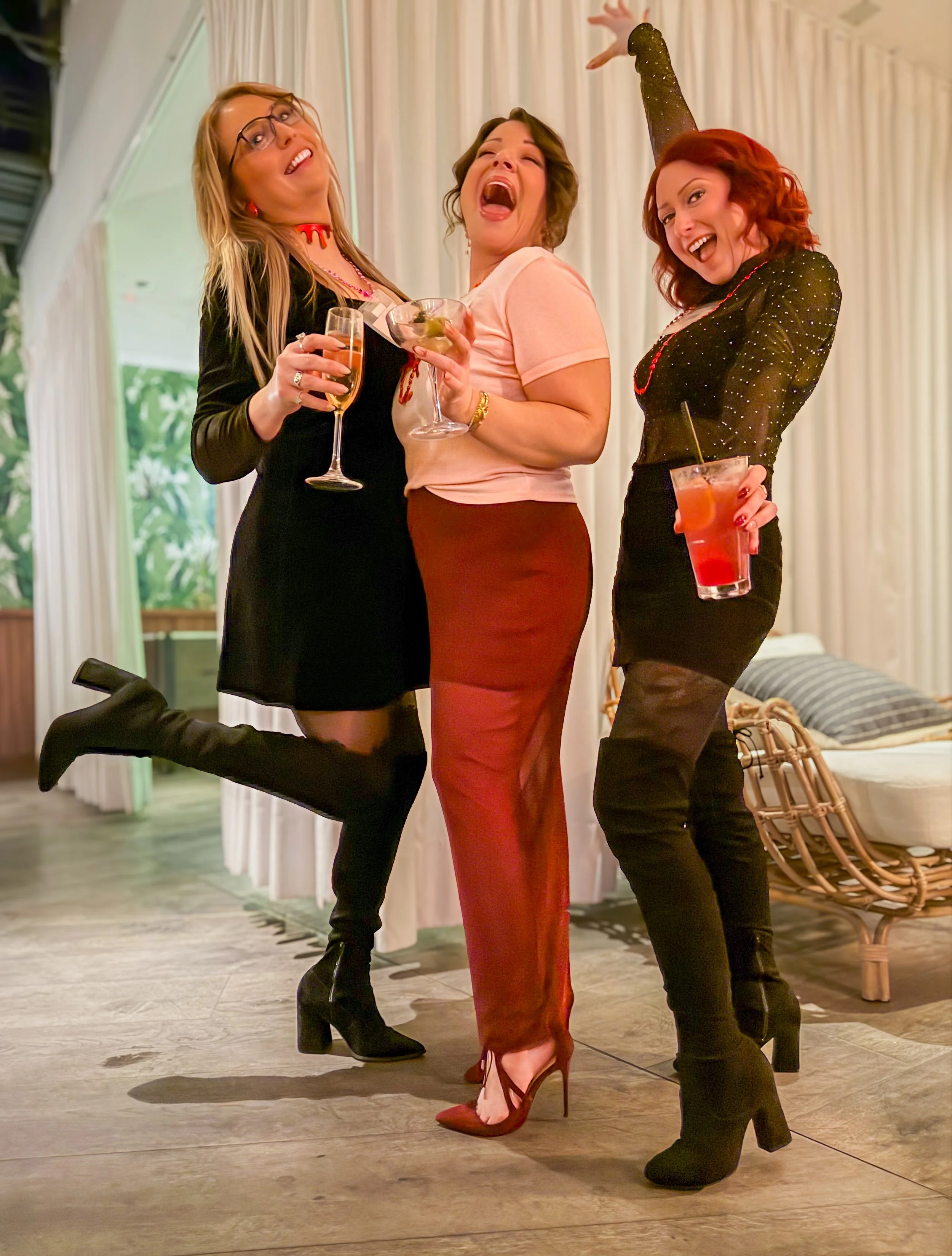Three women celebrating at a party, all holding drinks and dressed in party attire, with the woman in the middle laughing.