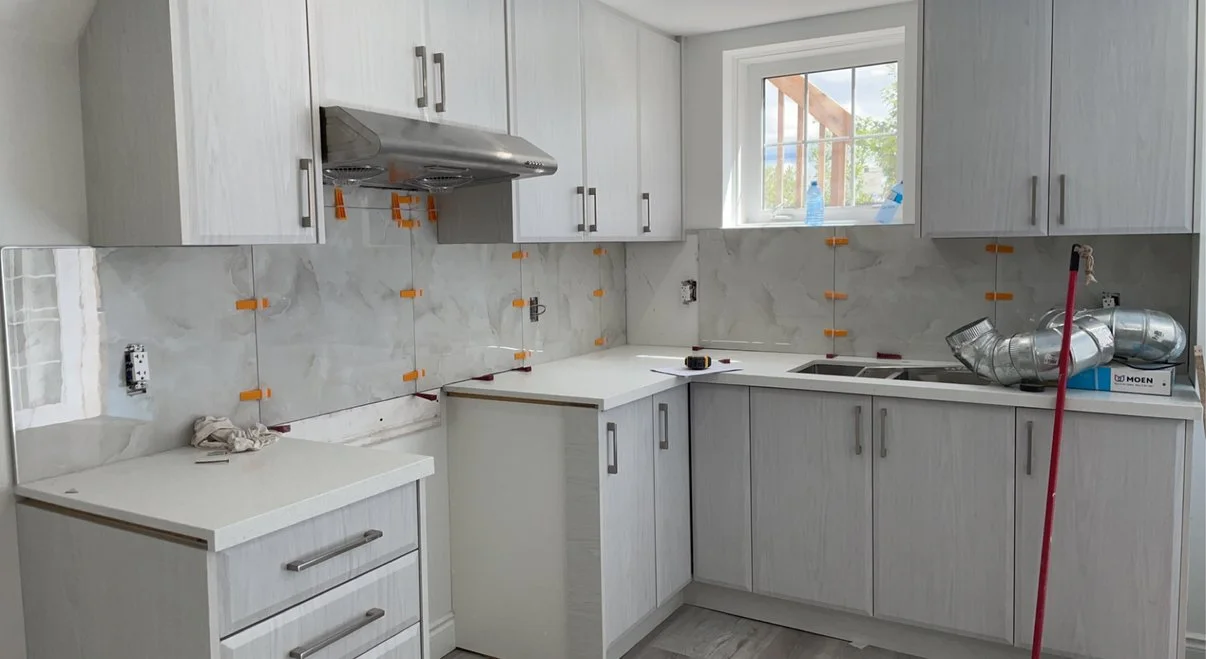Kitchen under renovation with white cabinets and marble backsplash, window above sink, construction tools and materials, and a red mop handle leaning against the counter.