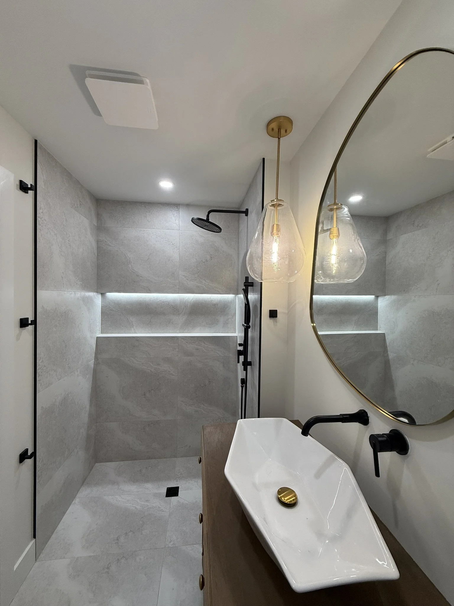 Modern bathroom with gray stone shower, black fixtures, white vessel sink with black faucet, oval mirror, and two hanging pendant lights.