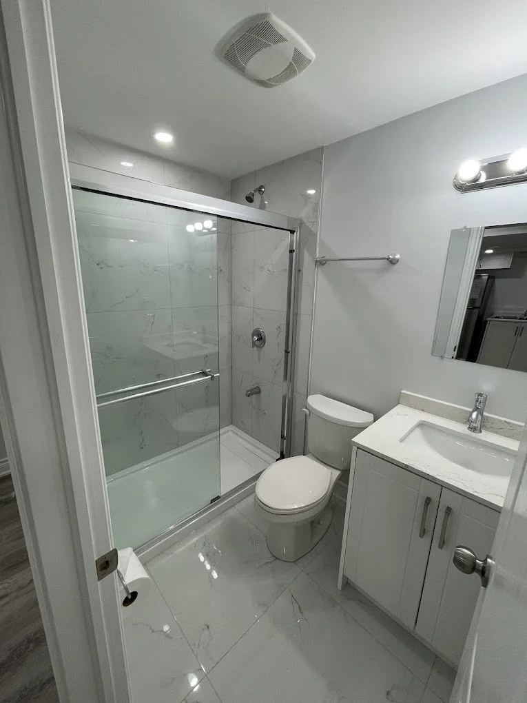 Small modern bathroom with a shower enclosed by glass sliding doors, a white toilet, and a white vanity with a sink. There is a mirror above the vanity and a light fixture with three bulbs. The bathroom has light-colored marble or tile flooring and l