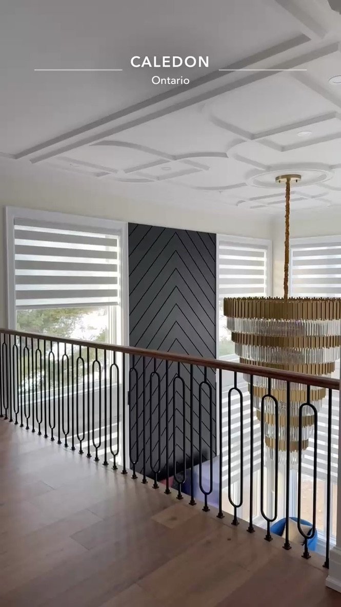 View of an interior staircase with a black decorative railing, a large chandelier hanging from the ceiling, windows with horizontal blinds, and dark wall paneling in a modern home in Caledon, Ontario.