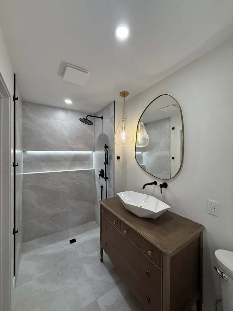 Modern bathroom with walk-in shower, white vessel sink on a wooden vanity, large oval mirror, pendant light, and neutral color scheme.