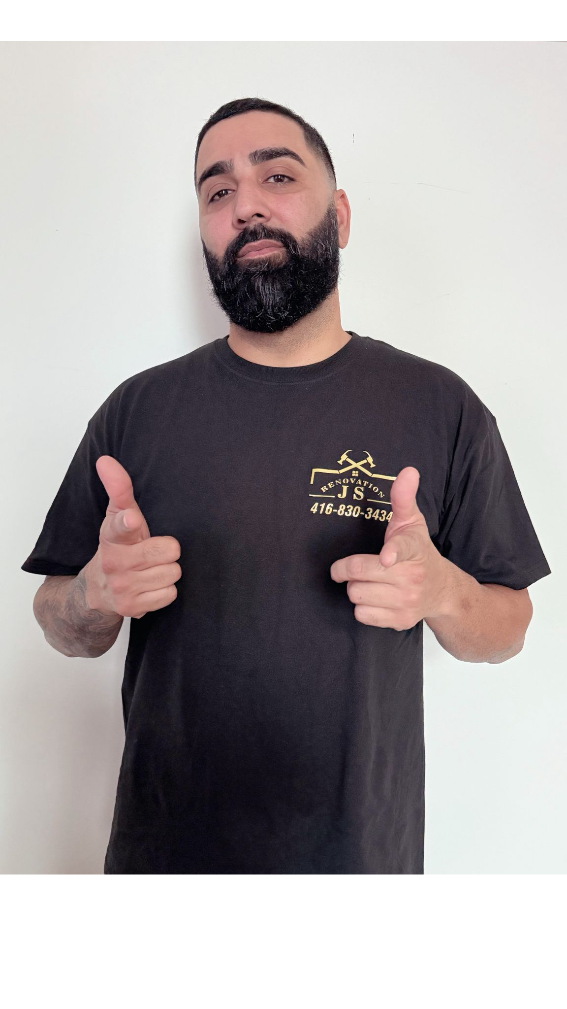 A man with a beard and mustache wearing a black T-shirt, pointing at himself with both hands, standing against a plain white wall.
