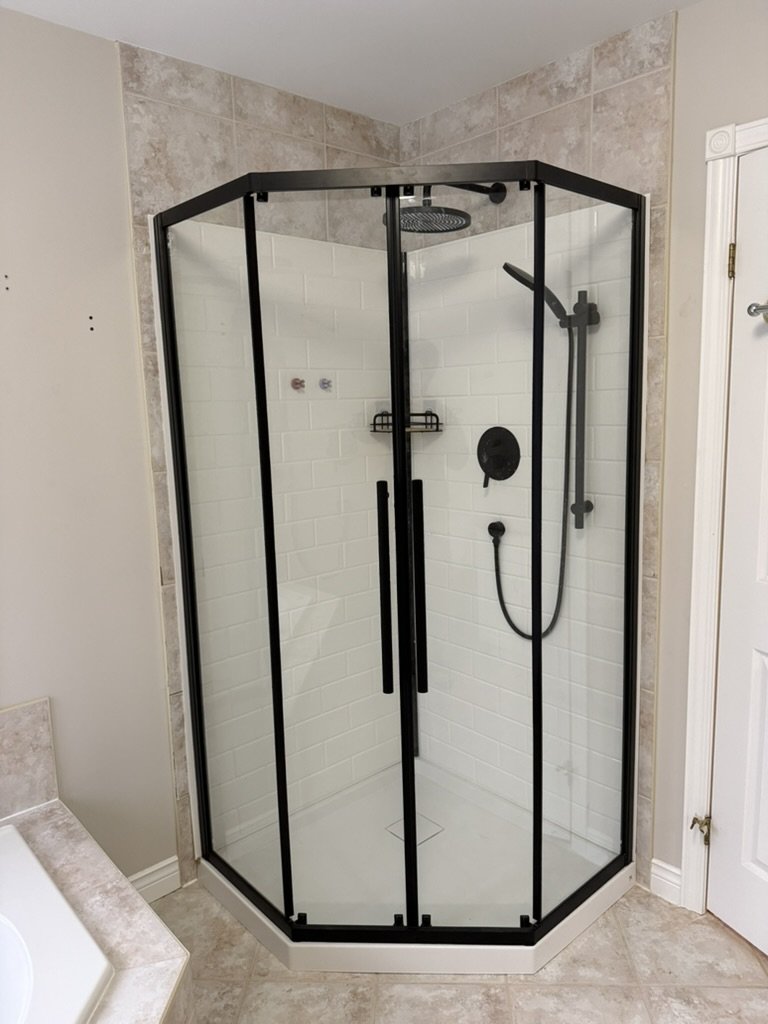 Black-framed glass shower enclosure with white subway tile interior, showerhead, handheld shower, and a corner shelf, in a bathroom with beige tiled floor and wall.