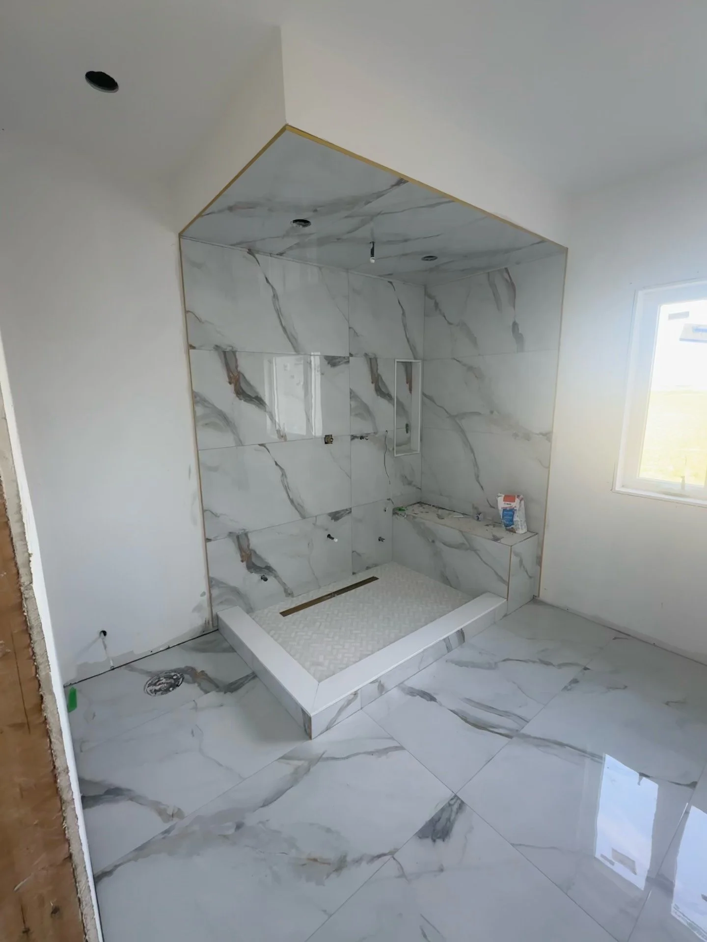 Bathroom under construction with a marble tiled shower area, a window, and partially completed wall and ceiling fixtures.