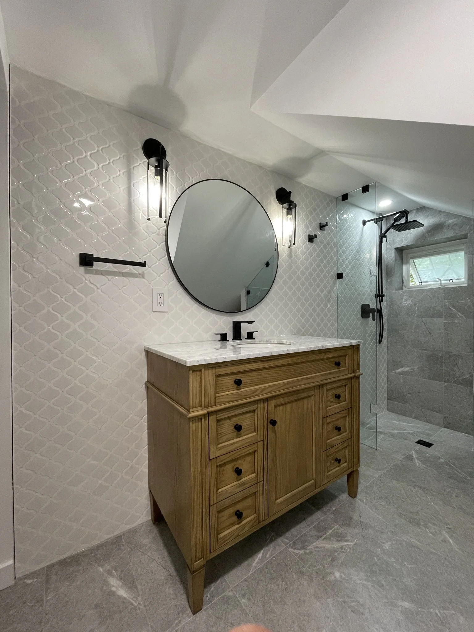 Modern bathroom with a wooden vanity, round mirror, black wall sconces, and a walk-in shower with a glass partition and a small window.
