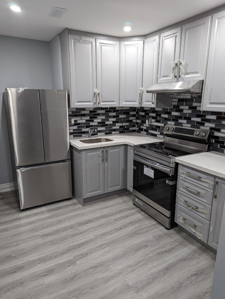 Modern kitchen with gray and white cabinets, stainless steel refrigerator and stove, black and gray tile backsplash, and light wood-look flooring.