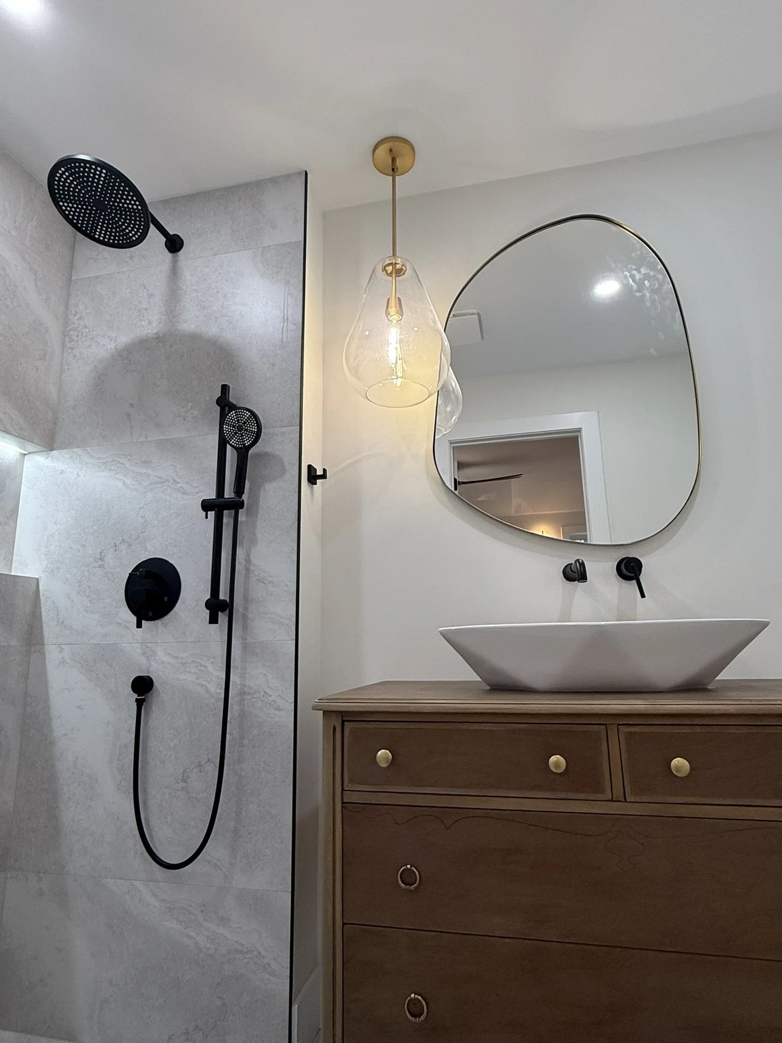 Modern bathroom with walk-in shower, black fixtures, a wooden vanity with a white vessel sink, a large irregularly shaped mirror, and a pendant light with glass shades.