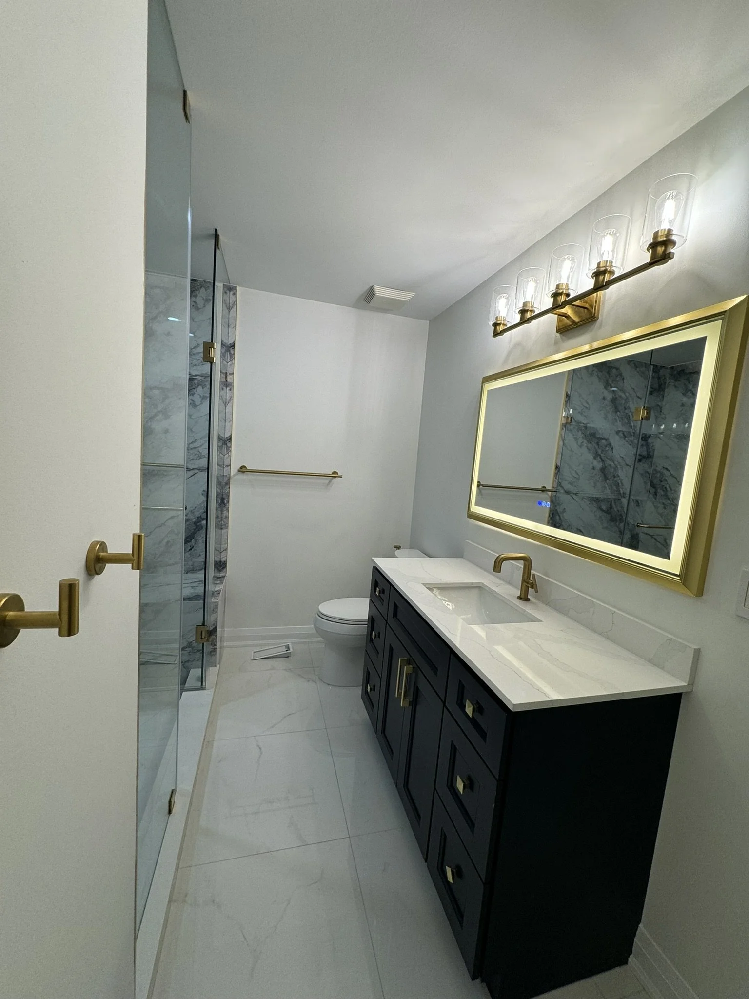 Modern bathroom with a black vanity with gold hardware, a white marble countertop with a built-in sink, a large rectangular framed mirror with gold trim, a wall-mounted gold light fixture with five glass shades, a toilet, a glass-enclosed shower with