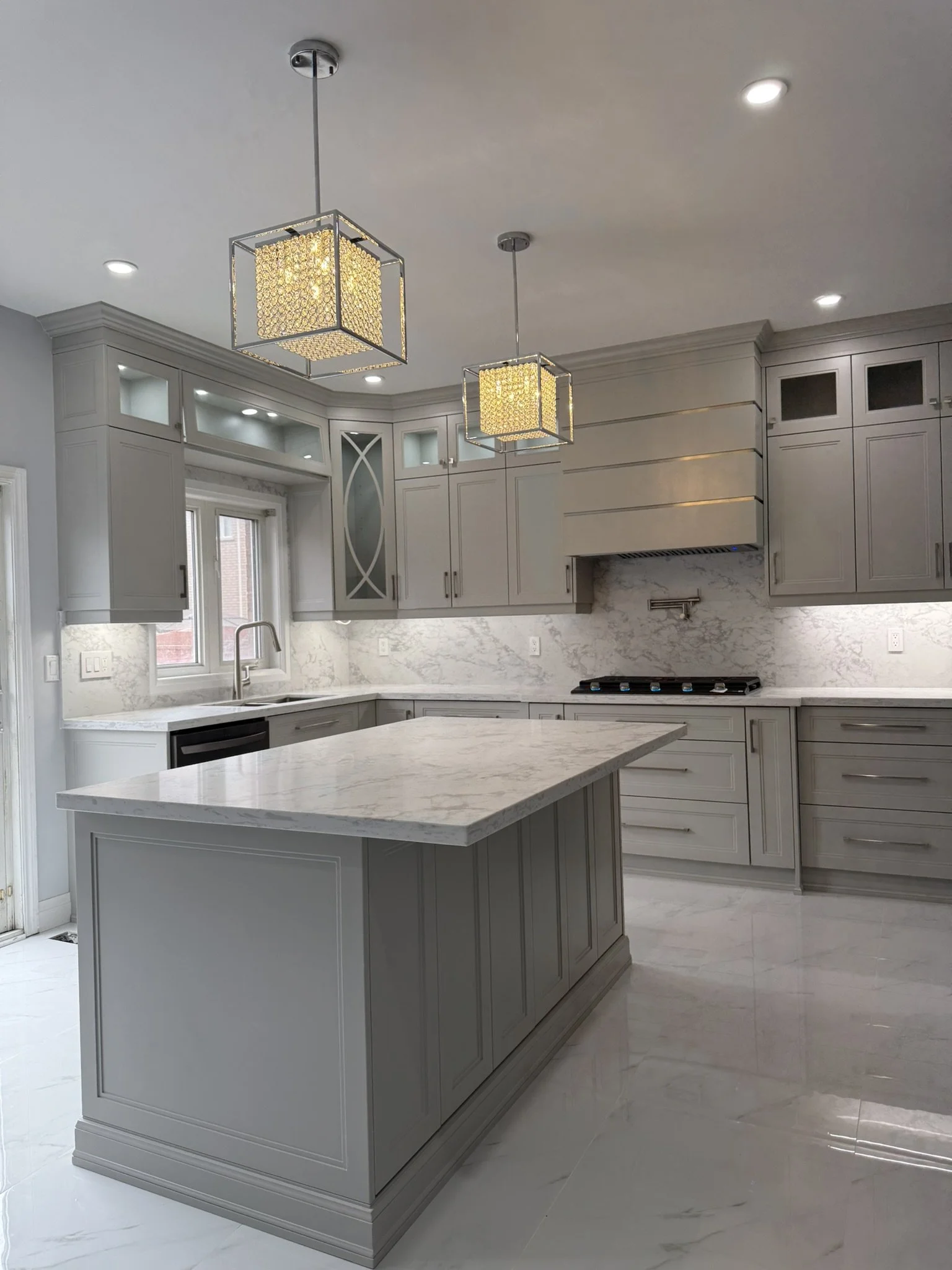 Modern kitchen with light gray cabinetry, marble countertops, and a marble backsplash, featuring an island, stainless steel appliances, and two square pendant lights hanging from the ceiling.
