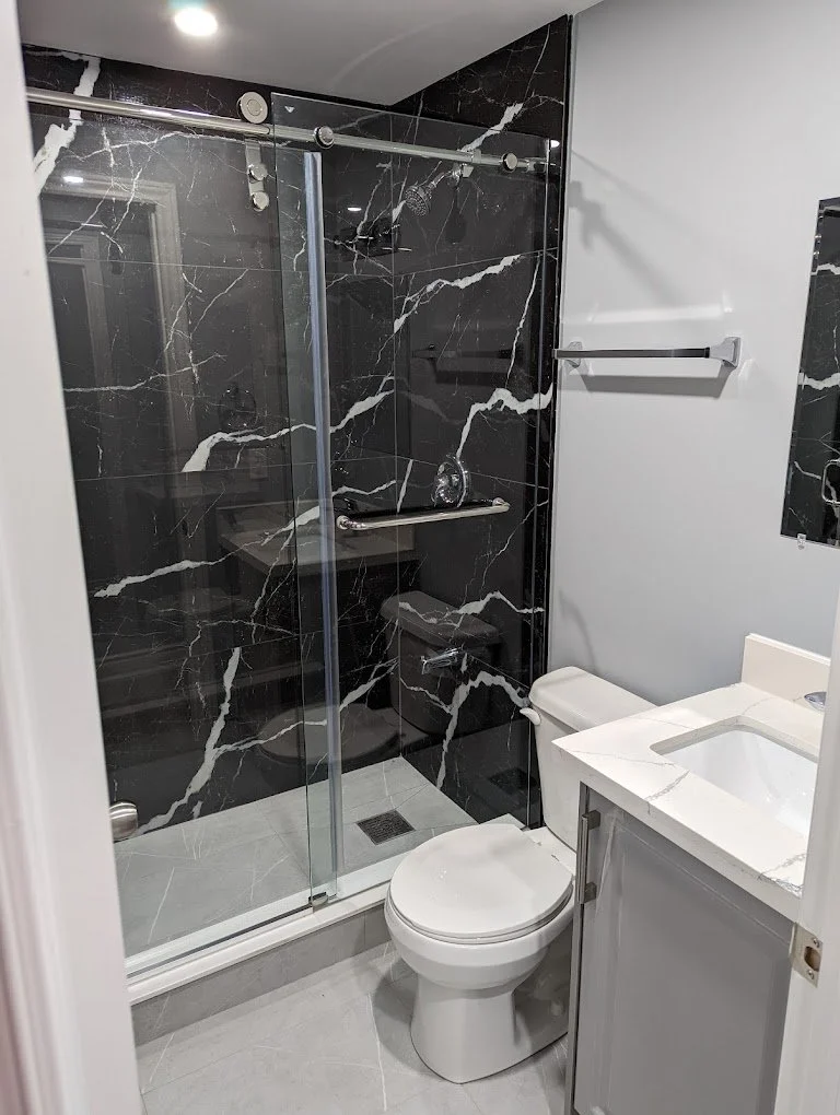 A compact modern bathroom with a black marble shower enclosure, a white toilet, and a white vanity with a sink. The shower has a glass door, a grab bar, and a small soap dish. There is a towel rack on the wall beside the shower.