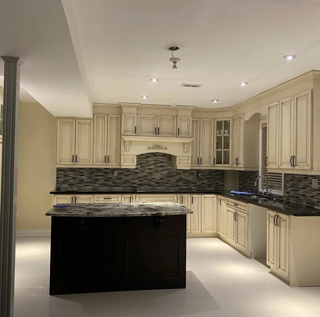 Modern kitchen with beige cabinets, black granite countertops, and a dark central island with a granite top. There is a backsplash with black, gray, and beige tiles, and a window above the sink. Recessed lighting illuminates the kitchen.