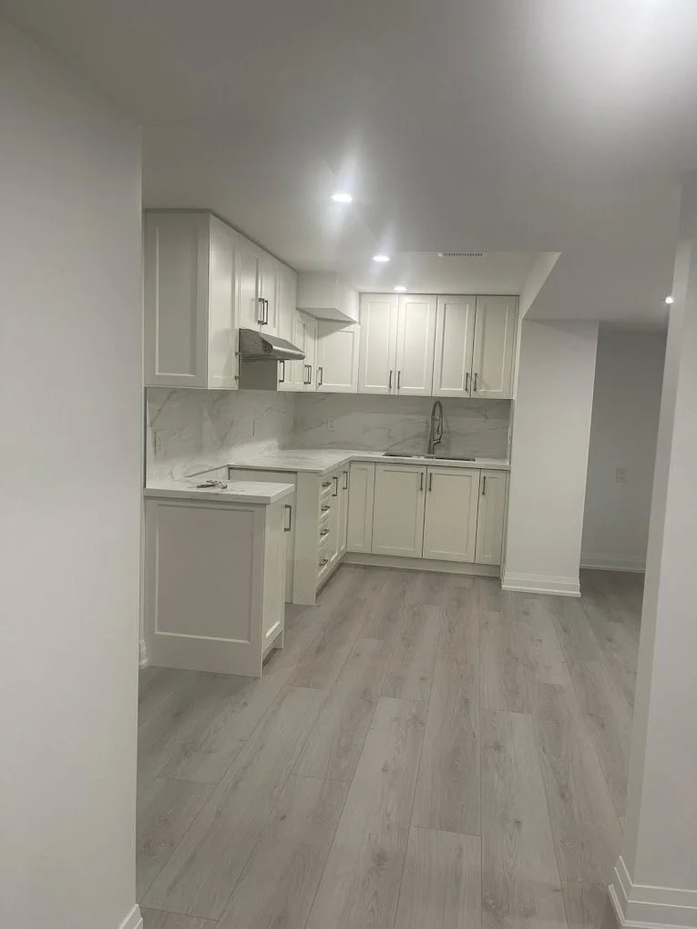 Empty modern kitchen with white cabinets, marble countertops, gray hardwood floors, and a stainless steel faucet.